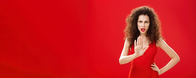 Aspetta ferma intensa donna adulta dall'aspetto serio e scontento con i capelli ricci in un elegante abito rosso