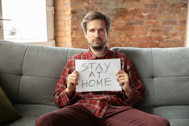 Holding banner STAY AT HOME sitting on sofa in room