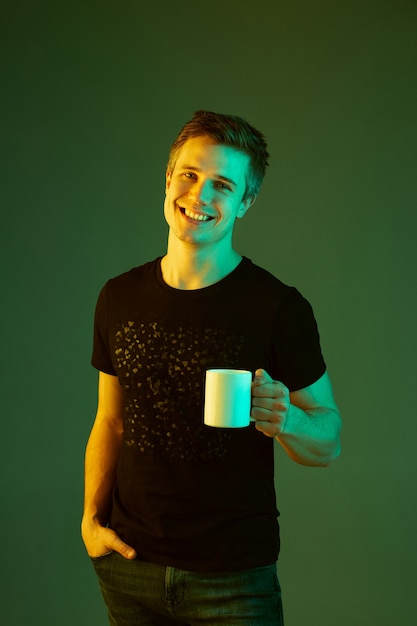 Holding cup and smiling. caucasian man's portrait isolated on green  background in neon light.