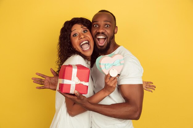 Holding giftboxes. Valentine's day celebration, happy african-american couple isolated on yellow studio background. 