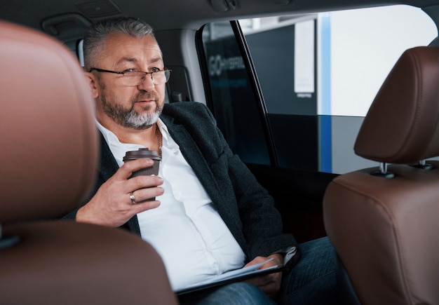Holding plastic cup of coffee. Paperwork on the back seat of the car. Senior businessman with documents