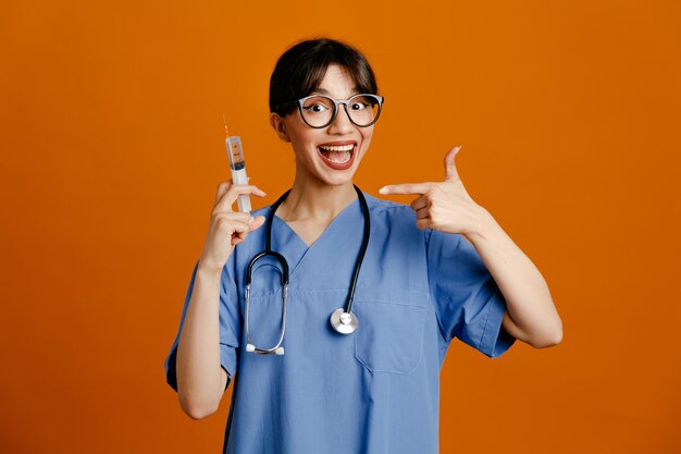 Holding and points at syringe young female doctor wearing uniform fith stethoscope isolated on orange background