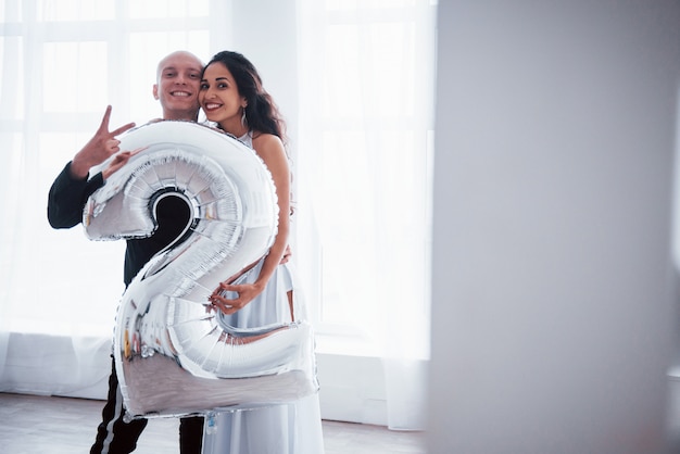 Holding silver colored balloon in the shape on number two. Young couple in luxury wear stands in the white room