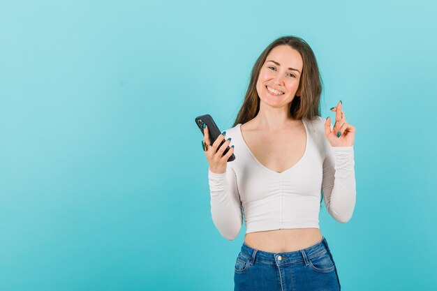 Holding smartphone girl is raising up other hand by crossing fingers on blue background