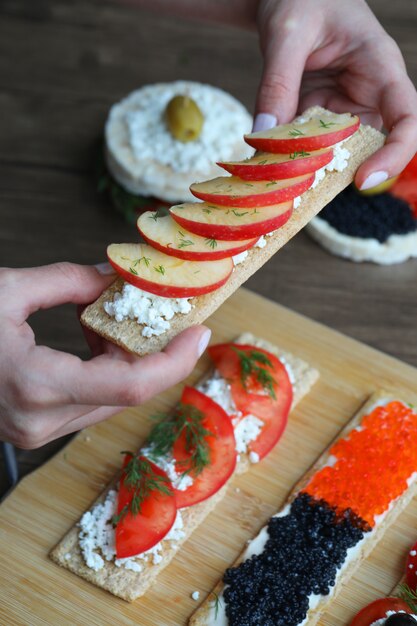 Holding a vegan cracker with sliced apple and ricotta cheese. Mixed crackers on the wooden board.