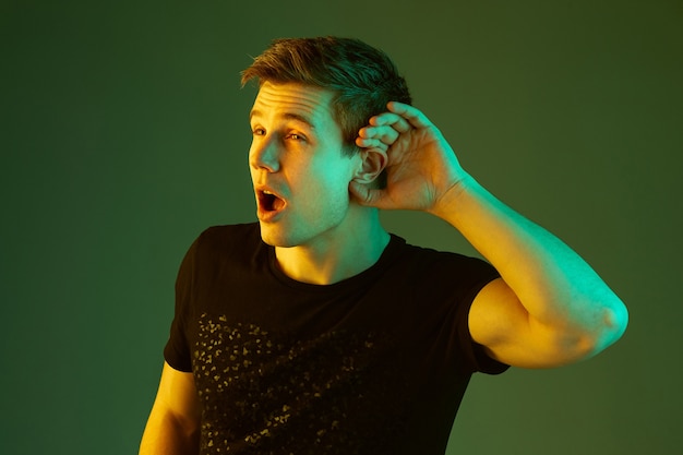 Holds palm on ear, listening. Caucasian man's portrait isolated on green studio background in neon light. Beautiful male model in black shirt. Concept of human emotions, facial expression, sales, ad.