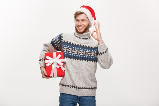 Holiday Concept Young beard man in sweater holding box and giving ok sign