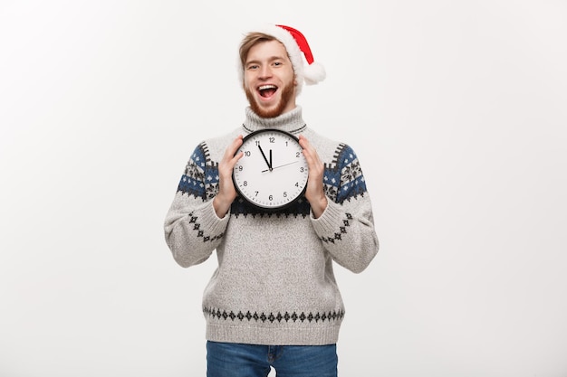 Holiday concept Young happy handsome beard man in sweater holding white clock over white studio background