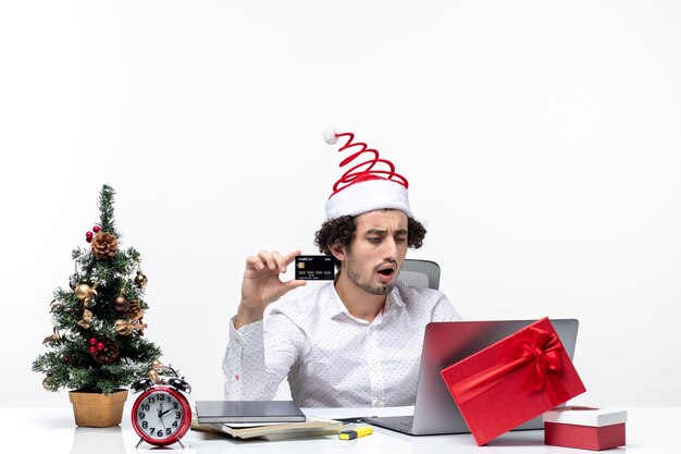 サンタクロースの帽子と白い背景の上のオフィスで一人でプロジェクトに取り組んでいる銀行カードを保持している驚きのビジネスパーソンと休日のお祭り気分