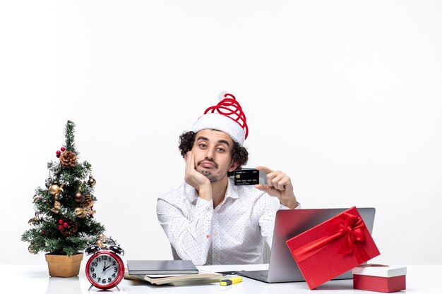 Празднично-праздничное настроение с молодым бородатым деловым человеком в шляпе санта-клауса, держащим банковскую карту и кладущим руку под подбородок в офисе