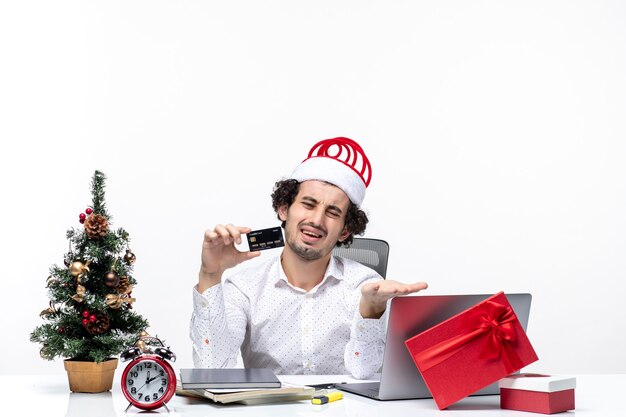 Umore festivo vacanza con giovane uomo d'affari arrabbiato stanco con cappello di babbo natale e tenendo la sua carta di credito in ufficio su sfondo bianco stock photo