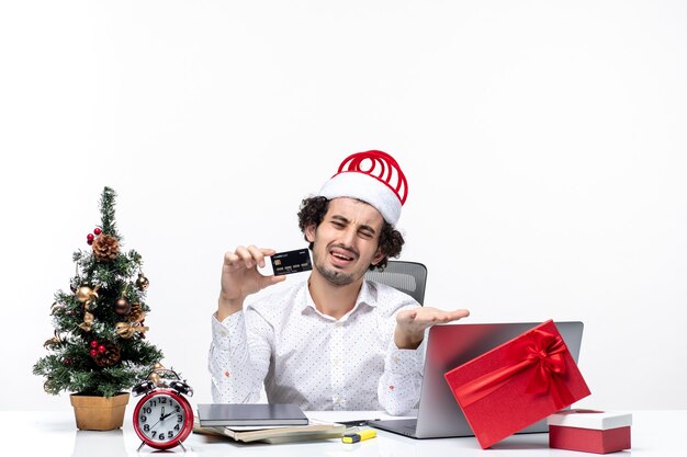 サンタクロースの帽子と白い背景の上のオフィスで彼の銀行カードを保持している若い疲れた怒っているビジネスパーソンと休日のお祭り気分ストックフォト