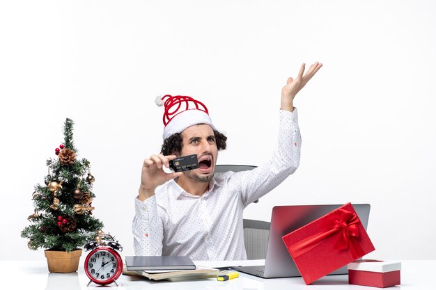 Holiday festive mood with young tired angry business person with santa claus hat and holding his bank card in the office on white background