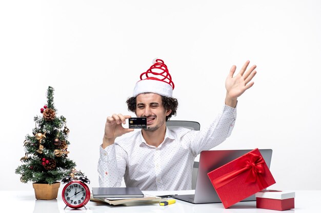 Holiday festive mood with young tired angry business person with santa claus hat and looking at his bank card in the office on white background