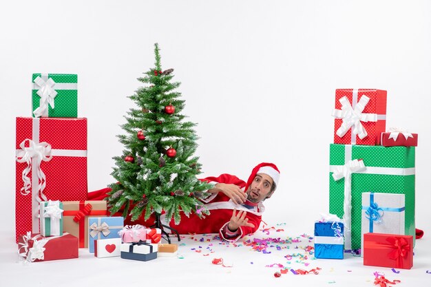 白い背景の上の贈り物の近くのクリスマスツリーの後ろに横たわっているサンタクロースと休日の気分