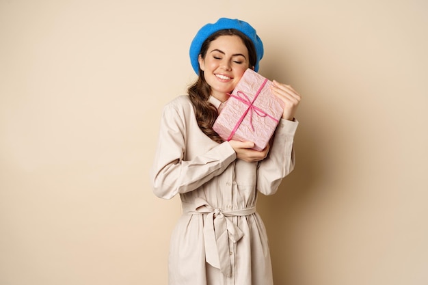 Бесплатное фото Концепция праздников и подарков. красивая девушка получает подарочную коробку и выглядит счастливой, держит розовый завернутый подарок с радостным выражением лица, бежевый фон