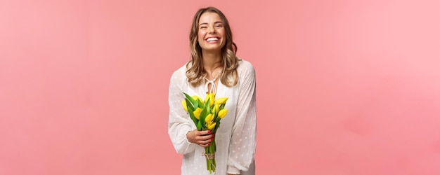 Holidays beauty and spring concept Carefree happy attractive blond girl in white dress holding yellow tulips and laughing with pleased expression standing pink background have romantic date