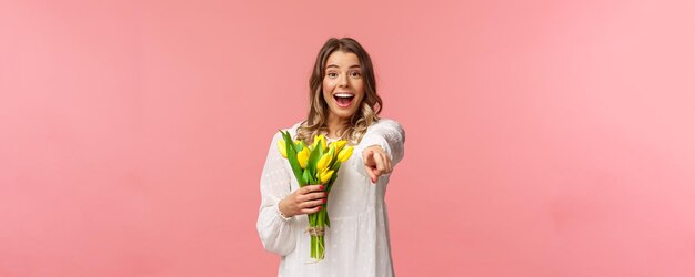 休日の美しさと春のコンセプトカメラに指を指している黄色いチューリップの花束を保持している白いドレスを着た興奮した幸せで明るい若いブロンドの女の子の肖像画驚いたピンクの背景