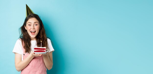 Holidays and celebration cheerful birthday girl in party hat holding bday cake and smiling making wi