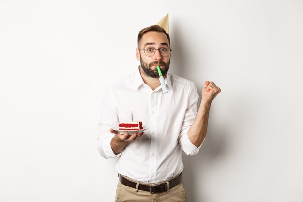 Holidays and celebration. Cheerful man enjoying birthday, blowing party whistle and holding bday cake 