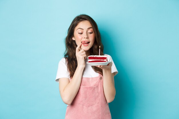 休日やお祝い。青い背景に夢のように立って、それを噛む誘惑でおいしいケーキを見ているかわいい誕生日の女の子。