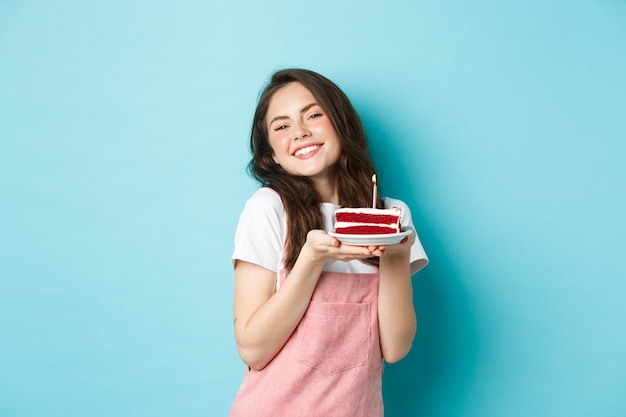 休日やお祝い。彼女の誕生日を祝って、ケーキとプレートを保持し、陽気な笑顔、祝って、青い背景の上に立っているかわいい魅力的な女の子。