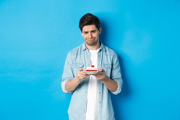 Holidays and celebration. Disappointed and sad man looking at birthday cake, grimacing upset, standing against blue background