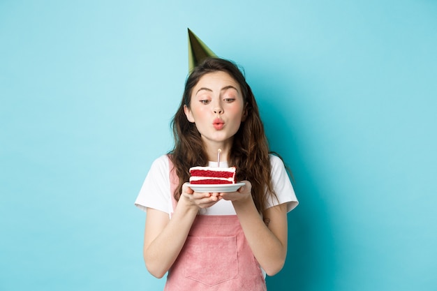 Праздники и торжества. Взволнованная женщина празднует день рождения, задувая свечу на торте, носит праздничный торт и веселится, стоя на синем фоне.