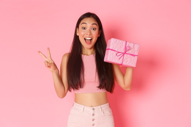 Holidays, celebration and lifestyle concept. Smiling kawaii asian girl showing wrapped gift and peace gesture, likes giving presents, standing over pink background. Copy space