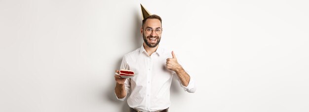 Holidays and celebration satisfied man enjoying bday party holding birthday cake and showing thumb u