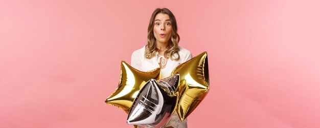 Holidays celebration and women concept Portrait of surprised happy attractive blond girl in white lovely dress fold lips say wow being impressed with friends made bday party hold balloons