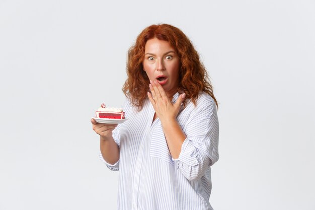 휴일, 감정 및 라이프 스타일 개념. 충격을 받고 걱정되는 중년의 빨간 머리 여자가 입을 벌리고 헐떡이며 케이크를 들고 걱정하는 표정으로 얼마나 많은 칼로리가 있는지 들어보십시오.