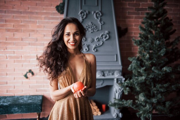 Holidays is coming. Pretty brunette holding ball shaped candle while stands indoor near the fireplace and Christmas tree