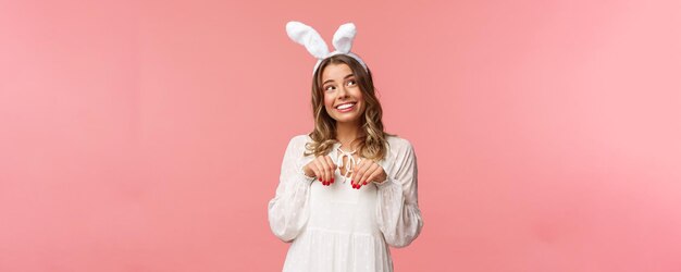 Holidays spring and party concept Cute romantic young blond girl imitating bunny wear white dress lovely rabbit ears make handpaws near chest and look dreamy up with daydreaming smile