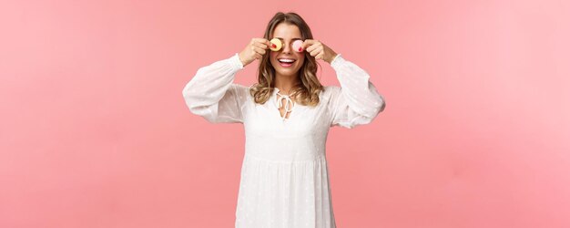 Holidays spring and party concept Portrait of carefree funny and cute blond girl in white dress holding two macarons on eyes and smiling happy like eating tasty desserts pink background