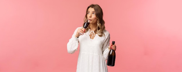 Holidays spring and party concept Portrait of carefree independent cute blond woman sipping wine from glass holding bottle and tasting drink look up celebrating with friends pink background