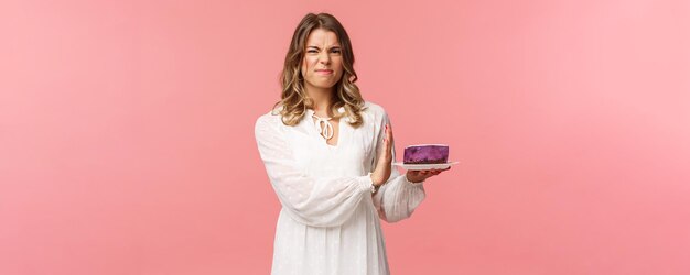 Holidays spring and party concept Portrait of dissatisfied and displeased blond girl in white dress rejecting declining eat tasteless cake grimacing and show stop sign at dessert pink background