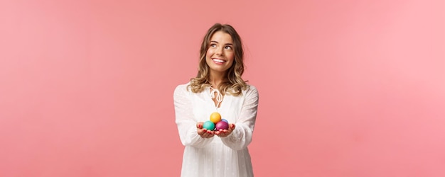 Vacanze primavera e concetto di festa ritratto di sognante bella ragazza bionda in abito bianco con uova dipinte che celebrano il giorno santo ortodosso di pasqua sorridente e distogliendo lo sguardo