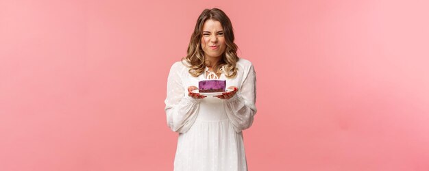 Holidays spring and party concept Portrait of excited and pleased blond female in white dress holding piece of delicious cake and grimacing satisfied cant wait to have bite of tasty dessert