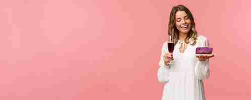 Free photo holidays spring and party concept portrait of tender elegant young blond woman holding glass of wine and cake with lit candle smiling pleased celebrating birthday pink background