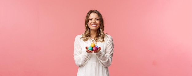 Vacanze primavera e concetto di festa ritratto di tenera bella ragazza bionda in abito bianco con uova dipinte di pasqua che celebrano il giorno ortodosso sorridente allegro condivisione positività sfondo rosa