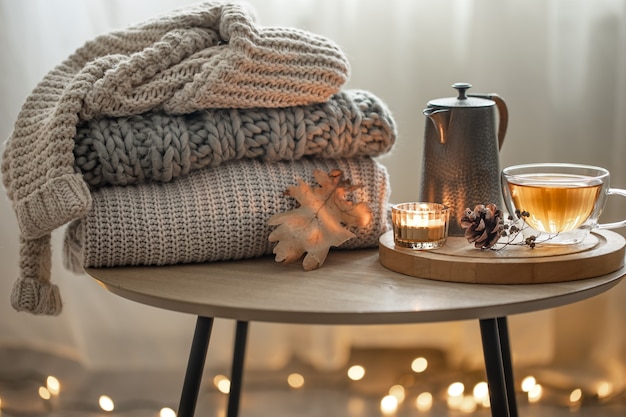 Домашняя осенняя композиция с чаем и вязанными свитерами в интерьере комнаты на размытом фоне с гирляндой.