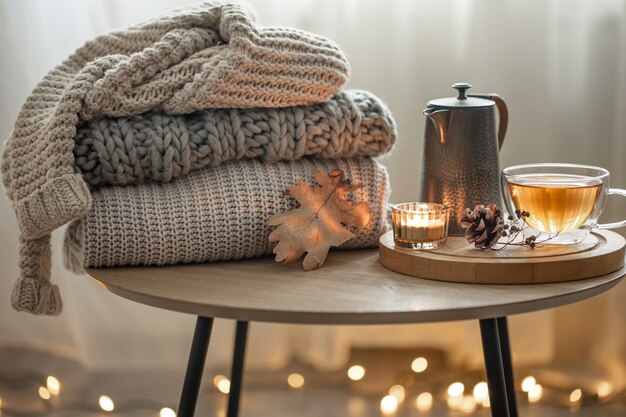 Home autumn composition with tea and knitted sweaters in the interior of the room, on a blurred background with a garland.