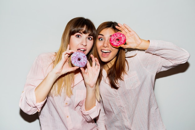 Home pajamas party. Flash portrait of two funny women posing with donuts. Surprise face.