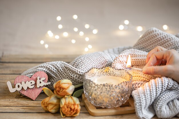 Home romantic still life with burning candle, decor, fresh flowers and knitted element on blurred background with bokeh.