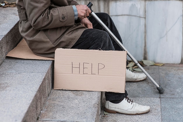 Homeless man on stairs with cane and help sign