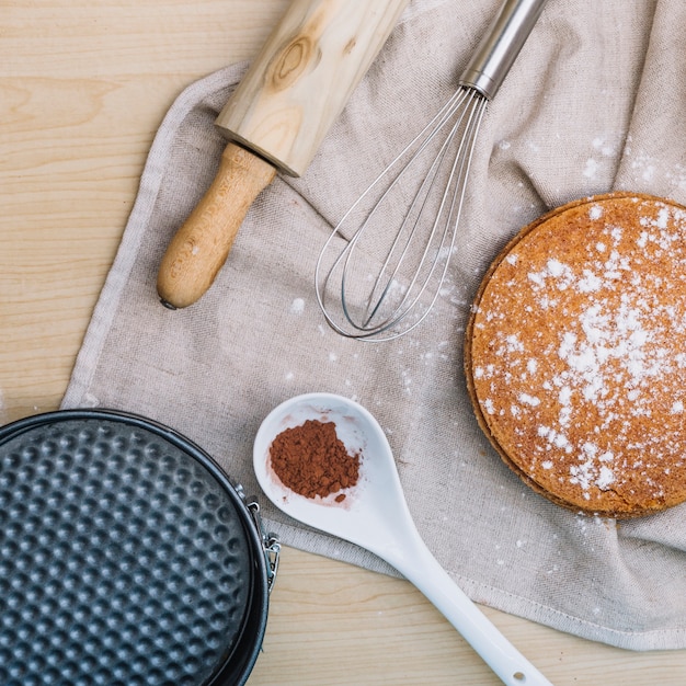 Free photo homemade cake base with chocolate powder; rolling pin; whisk and baking container on table