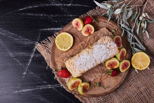 Homemade cake surrounded by various of fruits on round wooden platter. 