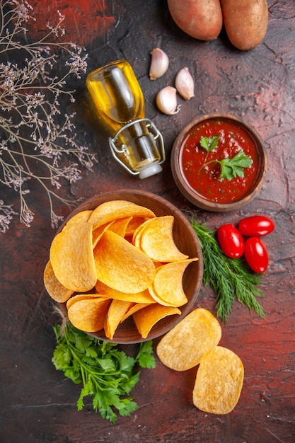 Homemade delicious potato crispy chips in a small brown bowl potatoes oil bottle green tomatoes garlic and ketchup on dark table