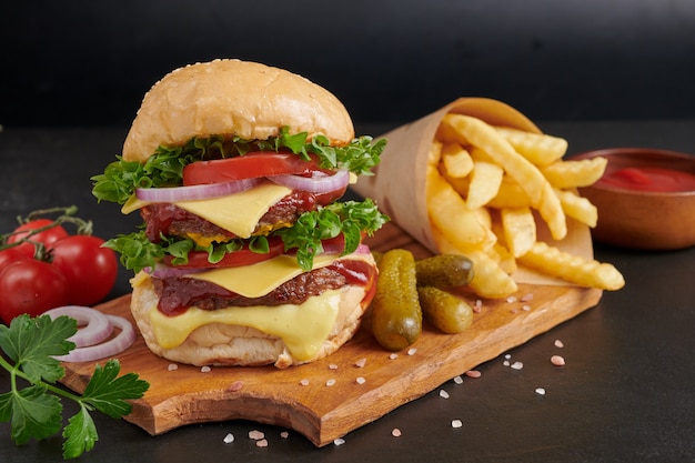 Домашний гамбургер или гамбургер со свежими овощами и сырным салатом и майонезом, картофель фри на кусочках коричневой бумаги на черном каменном столе. концепция быстрого питания и нездоровой пищи